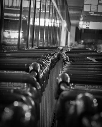 A monochrome image of a row of shopping carts lined up in a supermarket or store environment. The carts are tightly packed together, and the image is captured from a low angle, creating a sense of depth. The lighting is dim, with reflections on the metal surfaces, giving the scene an industrial feel.