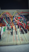A breadboard setup with numerous multi-colored connecting wires and electronic components, including resistors and LEDs. A computer monitor is visible in the background.