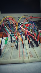 A breadboard setup with numerous multi-colored connecting wires and electronic components, including resistors and LEDs. A computer monitor is visible in the background.