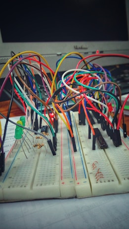 A breadboard setup with numerous multi-colored connecting wires and electronic components, including resistors and LEDs. A computer monitor is visible in the background.
