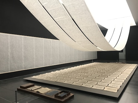 An art installation features large scrolls of text hanging from the ceiling, creating an arch-like form over a series of open books laid out in a grid on a platform. The room walls are adorned with text panels. The space is minimalistic with a monochrome palette, giving a sense of calm and contemplation.