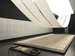 An art installation features large scrolls of text hanging from the ceiling, creating an arch-like form over a series of open books laid out in a grid on a platform. The room walls are adorned with text panels. The space is minimalistic with a monochrome palette, giving a sense of calm and contemplation.