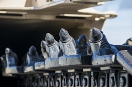 Close-up view of a series of metal components with a rugged, industrial appearance. The elements have a cylindrical tip, structured in a row along a mechanical part, likely part of heavy machinery such as a trencher or drilling equipment. The background is softly blurred, focusing attention on the sharp and metallic texture of the components.