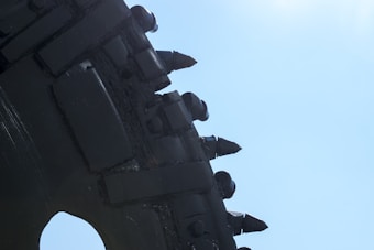 A close-up view of a large industrial drill or cutting machine component with metal teeth against a bright blue sky.