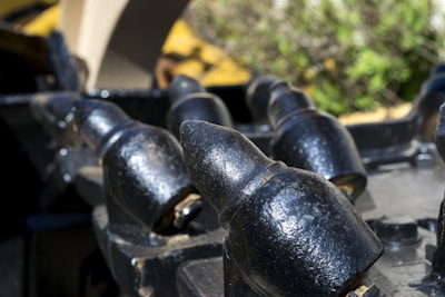 Close-up of heavy metal teeth or spikes, possibly part of industrial machinery, with a weathered black finish and some metal wear. The background is blurred, featuring green and yellow hues, likely from plants or foliage.