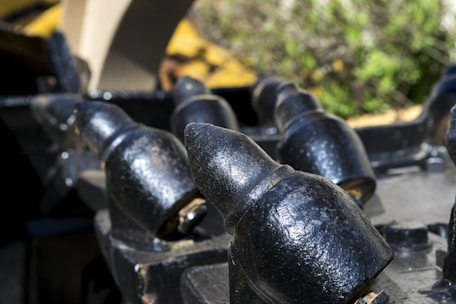 Close-up of heavy metal teeth or spikes, possibly part of industrial machinery, with a weathered black finish and some metal wear. The background is blurred, featuring green and yellow hues, likely from plants or foliage.