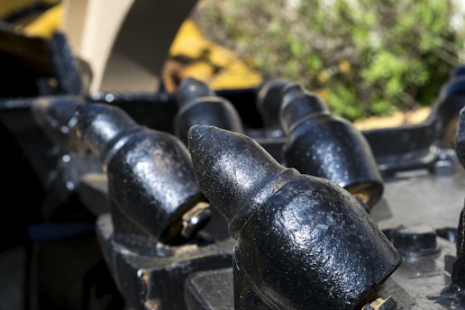 Close-up of heavy metal teeth or spikes, possibly part of industrial machinery, with a weathered black finish and some metal wear. The background is blurred, featuring green and yellow hues, likely from plants or foliage.