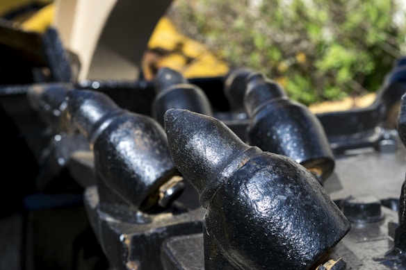 Close-up of heavy metal teeth or spikes, possibly part of industrial machinery, with a weathered black finish and some metal wear. The background is blurred, featuring green and yellow hues, likely from plants or foliage.