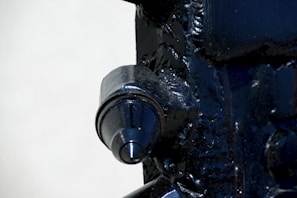 A close-up view of a metallic machine part with a prominent cylindrical feature. The surface is dark and glossy, with visible welds and textural details on the surrounding metal.