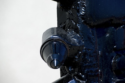 A close-up view of a metallic machine part with a prominent cylindrical feature. The surface is dark and glossy, with visible welds and textural details on the surrounding metal.