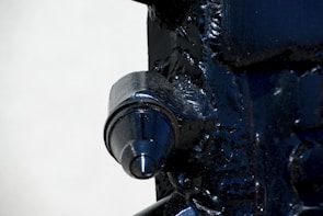 A close-up view of a metallic machine part with a prominent cylindrical feature. The surface is dark and glossy, with visible welds and textural details on the surrounding metal.