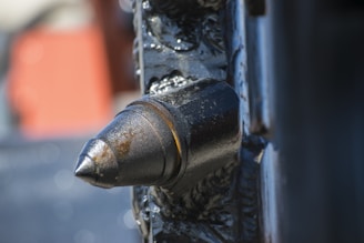 A close-up view of a black metallic structure featuring a prominent cylindrical and pointed protrusion, possibly part of an industrial machine or tool. The surface of the metal appears weathered or worn, with visible scratches and texture details. The background is blurred, highlighting the object in focus.