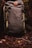 A sturdy, olive green backpack with a leather patch featuring a logo. It rests against a backdrop of forest floor, surrounded by earthy elements such as twigs, leaves, and moss.