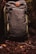 A sturdy, olive green backpack with a leather patch featuring a logo. It rests against a backdrop of forest floor, surrounded by earthy elements such as twigs, leaves, and moss.