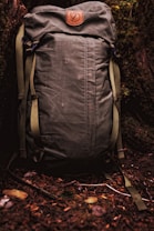 A sturdy, olive green backpack with a leather patch featuring a logo. It rests against a backdrop of forest floor, surrounded by earthy elements such as twigs, leaves, and moss.