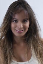 A young woman with long, dark brown hair and subtle highlights is smiling. She is wearing a white sleeveless top and has a natural makeup look with glossy lips. The background is plain white.