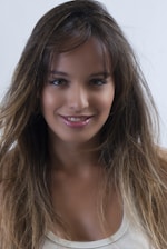 A young woman with long, dark brown hair and subtle highlights is smiling. She is wearing a white sleeveless top and has a natural makeup look with glossy lips. The background is plain white.