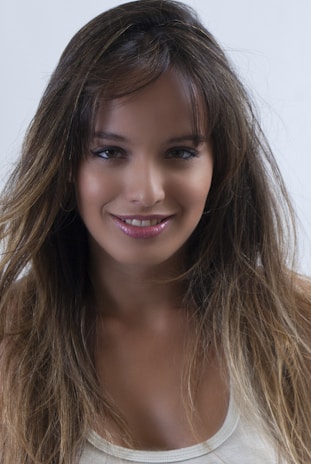 A young woman with long, dark brown hair and subtle highlights is smiling. She is wearing a white sleeveless top and has a natural makeup look with glossy lips. The background is plain white.
