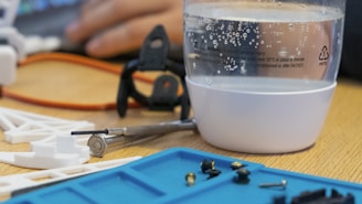A desk setup with a clear plastic cup filled with water, displaying bubbles on the surface. The cup has printed warnings and recycling symbols. In the foreground, there are various small mechanical components and tools on a blue mat. An orange cable loops around, and a blurred hand works in the background.