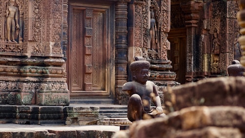 Intricately carved temple facade with detailed stone sculptures and ornate reliefs. A prominent statue of a seated figure positioned in the foreground adds depth and emphasis to the architectural marvel.