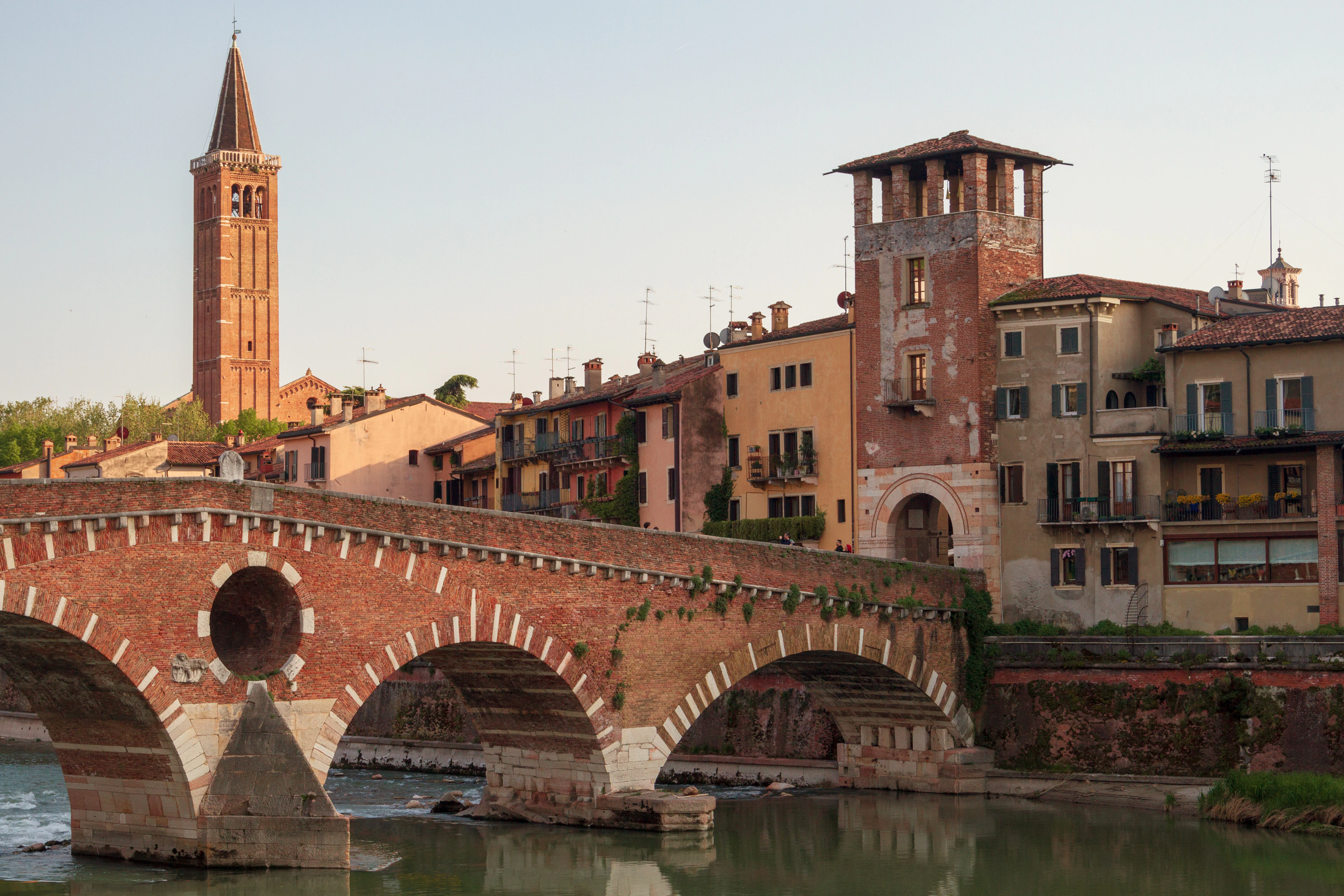 The arched stone Ponte Pietra crosses the calm Adige River at golden hour, bell towers rising behind it.