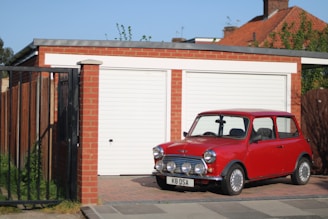 A classic red Mini Cooper is parked in front of a brick garage with white doors. The car's license plate reads 'K8 OSA'. The garage is part of a residential property with a black metal gate to the left and a wooden fence. The surrounding area appears suburban with visible rooftops and greenery in the background.
