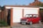 A classic red Mini Cooper is parked in front of a brick garage with white doors. The car's license plate reads 'K8 OSA'. The garage is part of a residential property with a black metal gate to the left and a wooden fence. The surrounding area appears suburban with visible rooftops and greenery in the background.