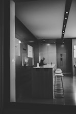 A person is sitting alone at a modern, minimalistic counter in what appears to be a technology store or showroom. The interior is sleek, with a row of high stools along the counter and large screens or displays on the walls. The lighting is subdued, contributing to the serene and somewhat isolated atmosphere of the space.