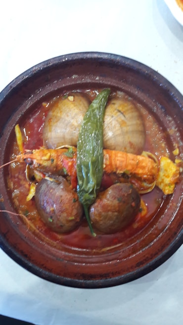 A rustic clay dish containing a seafood stew with large clams, a langoustine, potatoes, and a green chili pepper in a red sauce.