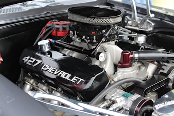 A close-up of a high-performance Chevrolet 427 engine with visible branding on the valve cover. The engine is equipped with a chrome air filter cover and several connected hoses and components, indicating a well-maintained and possibly restored condition.