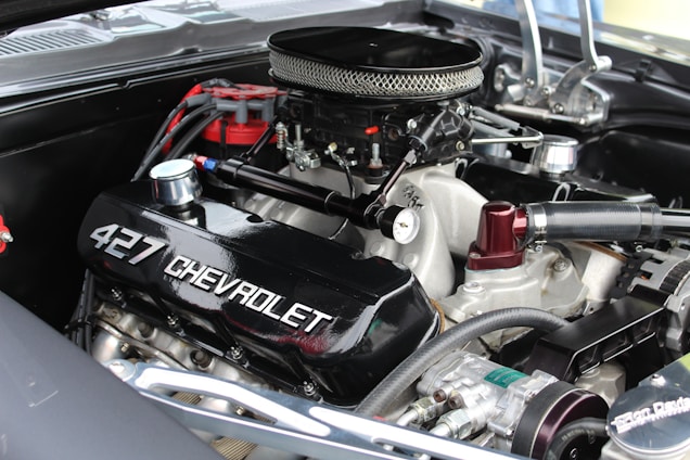 A close-up of a high-performance Chevrolet 427 engine with visible branding on the valve cover. The engine is equipped with a chrome air filter cover and several connected hoses and components, indicating a well-maintained and possibly restored condition.