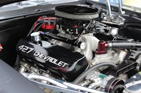 A close-up of a high-performance Chevrolet 427 engine with visible branding on the valve cover. The engine is equipped with a chrome air filter cover and several connected hoses and components, indicating a well-maintained and possibly restored condition.