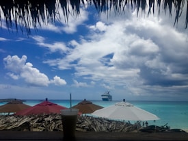A picturesque beach scene featuring a cruise ship in the distance on the vibrant blue ocean. Several brightly colored umbrellas are lined up along the sandy shore, with a straw-covered structure in the foreground providing shade. A tall glass with a straw is positioned in front, adding to the relaxed, tropical atmosphere. The sky is partly cloudy, with a mix of white and gray clouds contrasting against the deep blue.