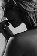 A grayscale portrait captures the side profile of a person with long hair. The focus is on their contemplative expression as they gently touch their lips with a hand. The lighting creates soft shadows, highlighting the texture of the skin and hair.