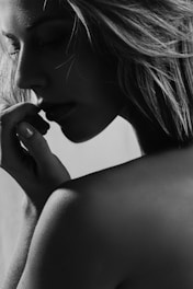 A grayscale portrait captures the side profile of a person with long hair. The focus is on their contemplative expression as they gently touch their lips with a hand. The lighting creates soft shadows, highlighting the texture of the skin and hair.