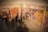 A gallery setting containing large, vibrant abstract paintings with circular and floral motifs. The artworks are predominantly in warm colors, and there are several people viewing the display, their reflections creating a layered effect. The scene feels dynamic due to the overlapping silhouettes of the viewers.