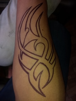 An arm with a large tribal tattoo in dark ink featuring a complex geometric pattern of sharp, interconnected lines and curves. The tattoo covers a significant portion of the forearm.