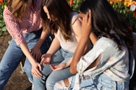 Three women sit closely together outdoors, engaged in conversation. They are wearing casual clothing, and their body language suggests they are sharing an intimate moment. The setting includes a garden with red and white tulips, indicating a bright and sunny day.