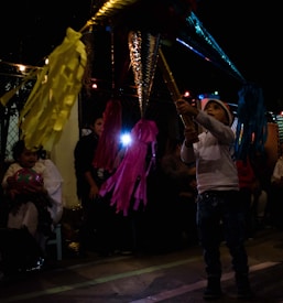 A child wearing a white sweater and a Santa hat is hitting a colorful piñata with bright streamers. The scene takes place in a dimly lit setting with hanging string lights and people sitting in the background. The atmosphere suggests a festive event or party, possibly a celebration with the piñata as a focal point.