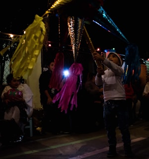 A child wearing a white sweater and a Santa hat is hitting a colorful piñata with bright streamers. The scene takes place in a dimly lit setting with hanging string lights and people sitting in the background. The atmosphere suggests a festive event or party, possibly a celebration with the piñata as a focal point.