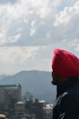 A person wearing a bright red turban and dark jacket gazes out over a scenic landscape featuring distant mountains and a partially cloudy sky. In the foreground, there's a tall building with a green roof and a cluster of trees.