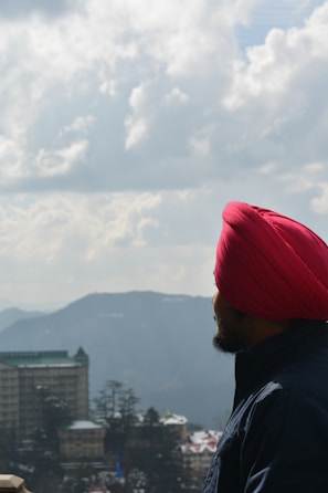 A person wearing a bright red turban and dark jacket gazes out over a scenic landscape featuring distant mountains and a partially cloudy sky. In the foreground, there's a tall building with a green roof and a cluster of trees.