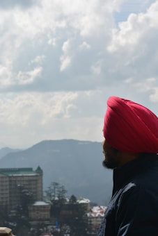 A person wearing a bright red turban and dark jacket gazes out over a scenic landscape featuring distant mountains and a partially cloudy sky. In the foreground, there's a tall building with a green roof and a cluster of trees.