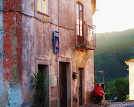 The image features an aged, rustic building with cracked walls and a small balcony. There's a street sign and a vibrant red scooter parked against the building. Lush greenery from a forested hillside is visible in the background under an early morning or late afternoon light, creating a tranquil setting.