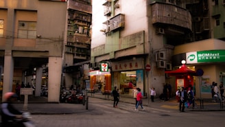 A bustling urban street corner features a mix of old and new architecture. A small convenience store is prominently displayed with its bright signage. Several people walk by, including some children and a person on a motorbike. The building facades are worn and weathered, typical of densely populated city areas.