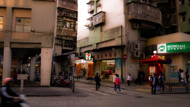 A bustling urban street corner features a mix of old and new architecture. A small convenience store is prominently displayed with its bright signage. Several people walk by, including some children and a person on a motorbike. The building facades are worn and weathered, typical of densely populated city areas.