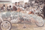 An outdoor gathering with a focus on custom motorcycles and a sizable crowd. Several unique motorcycles with extended handlebars are parked on the grassy field. People are dressed casually and appear to be enjoying the sunny day. The scene is lively with a blend of vibrant and neutral tones.