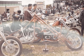 An outdoor gathering with a focus on custom motorcycles and a sizable crowd. Several unique motorcycles with extended handlebars are parked on the grassy field. People are dressed casually and appear to be enjoying the sunny day. The scene is lively with a blend of vibrant and neutral tones.