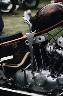 A close-up view of a custom motorcycle, showcasing intricate detailing on the gas tank and a unique, curved metal kickstand. The engine components are prominently displayed, highlighting the craftsmanship and vintage style.
