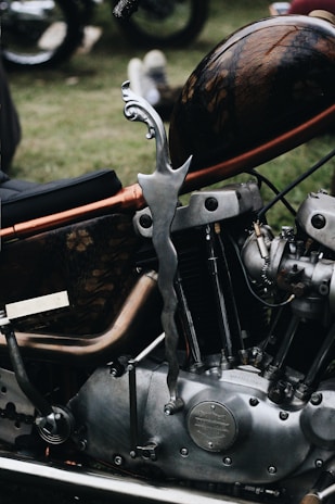A close-up view of a custom motorcycle, showcasing intricate detailing on the gas tank and a unique, curved metal kickstand. The engine components are prominently displayed, highlighting the craftsmanship and vintage style.